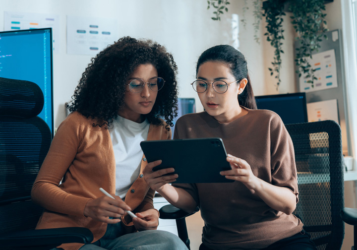 Two women sit in desk chairs looking at a tablet device. 