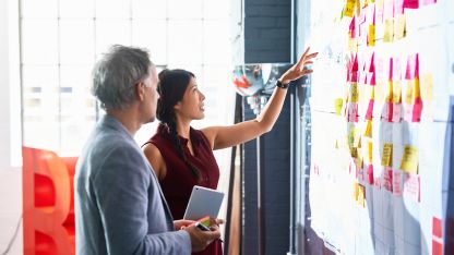 Two individuals engaged in discussion, standing before a whiteboard adorned with colorful sticky notes.