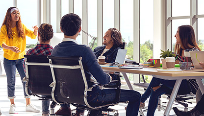 A female trainer wearing a yellow jumper is leading an energised class, in a sunny, modern office.